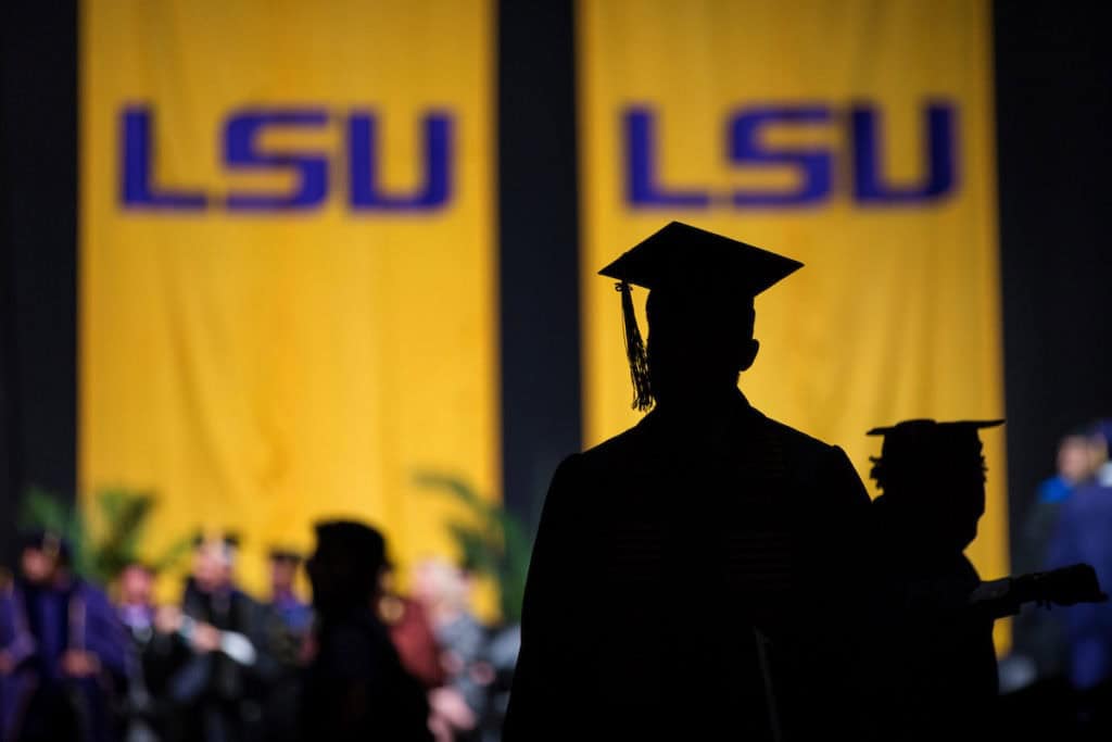 A silhouette of a graduate in cap and gown stands in front of two large yellow banners with "LSU" in purple letters.