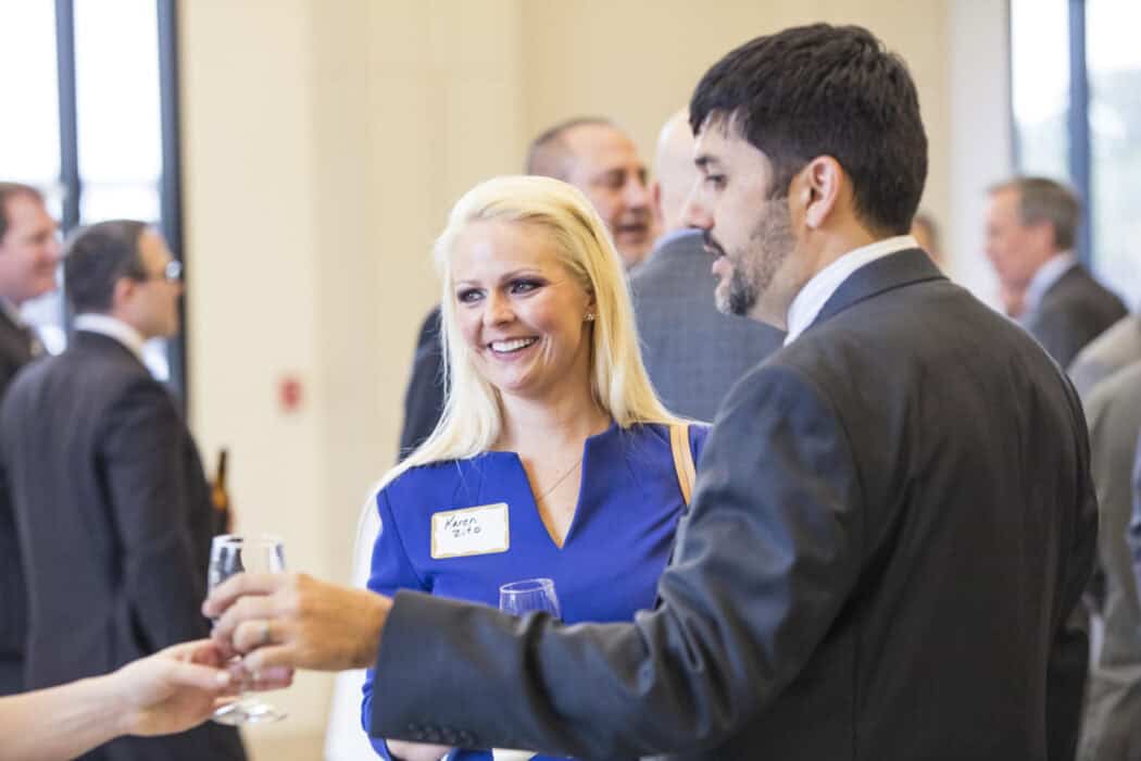 A woman and a man in business attire talk and hold drinks at a social event; other people are seen conversing in the background.