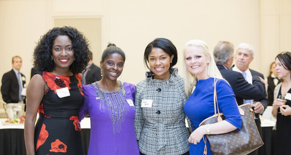 Four women pose together and smile at a formal indoor event, with other people mingling in the background.