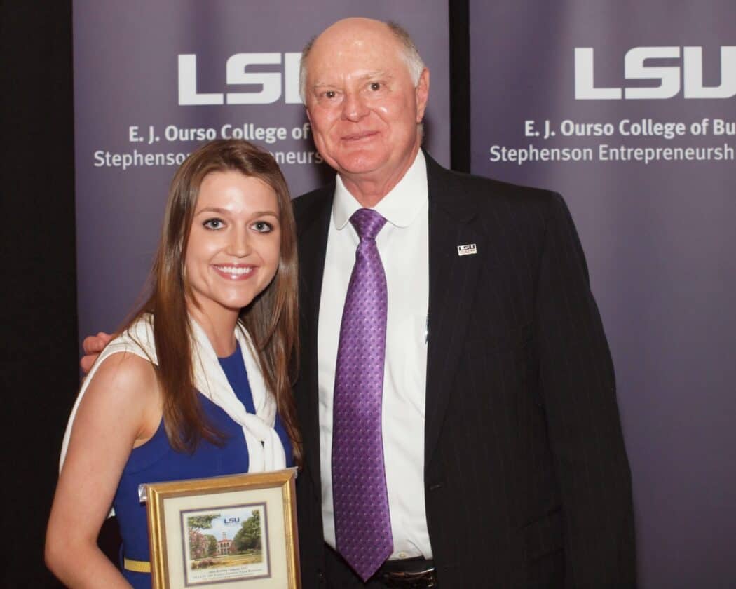 A young woman holding a framed certificate stands next to an older man in a suit, both posing in front of LSU College of Business banners.