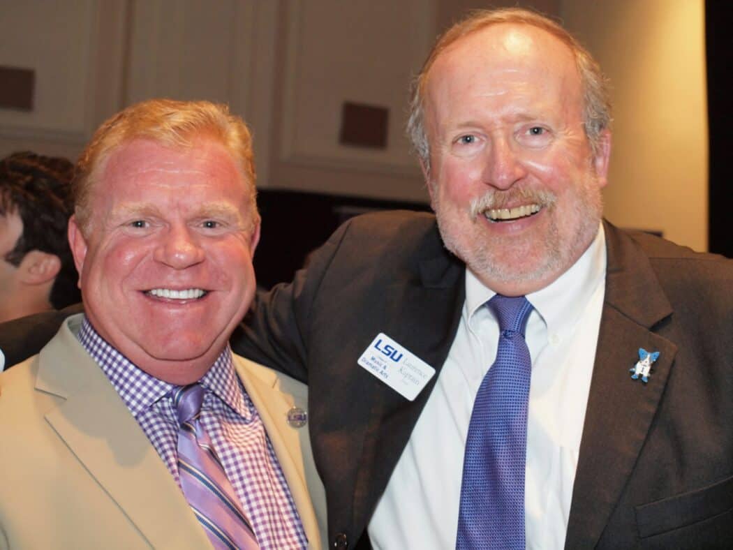 Two men in suits, one wearing a beige jacket and the other in a dark jacket, smile and pose together at an indoor event.