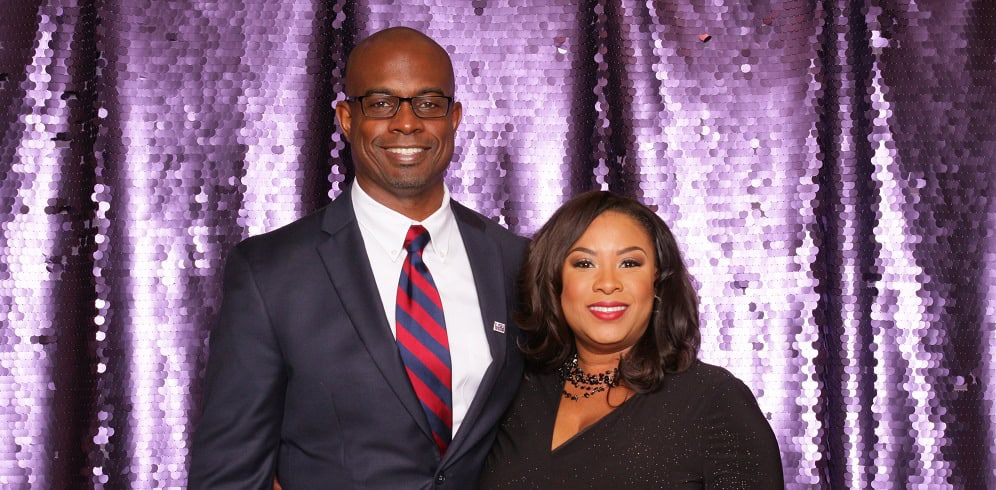 A man in a suit and a woman in a black dress stand together smiling in front of a purple sequin backdrop.