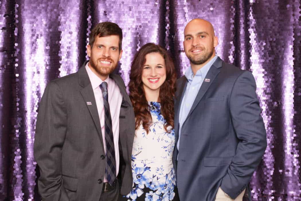Three people pose and smile in front of a purple sequin backdrop, dressed in business attire.