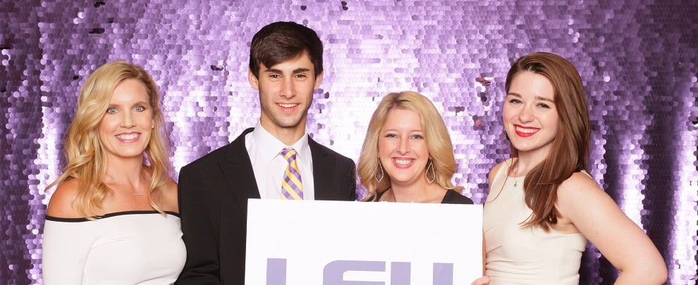 Four people in formal attire stand in front of a purple sequin backdrop, holding a large sign with "LSU" text.