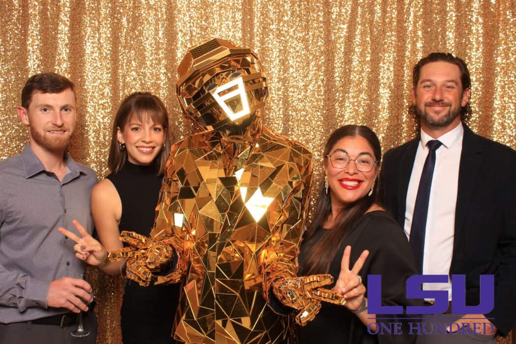 Five people pose in front of a gold sequin backdrop, including one in a gold geometric robot costume. LSU ONE HUNDRED logo is visible.