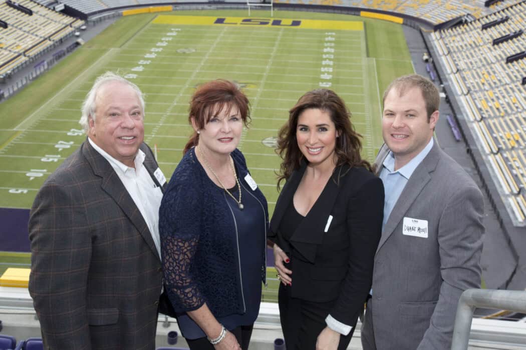 Four adults wearing business attire stand together in a stadium, with an empty LSU football field visible in the background.