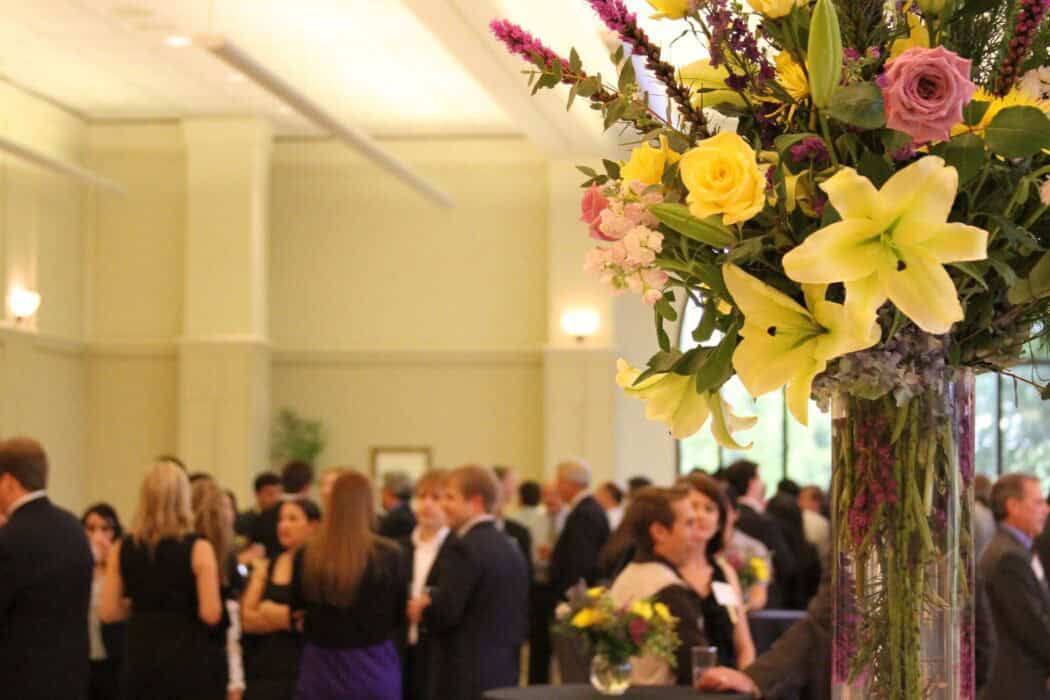 A large vase with yellow lilies and roses in focus; people in formal attire gather and mingle in a blurred background.