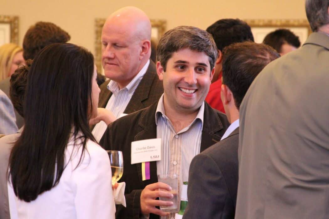 A group of people in business attire have a conversation at a social event. One person wears a visible name tag and holds a drink.