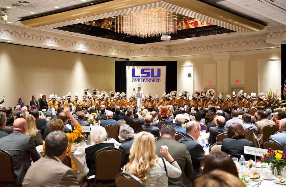 A large crowd sits at banquet tables watching a marching band perform onstage at an LSU One Hundred event in a ballroom.