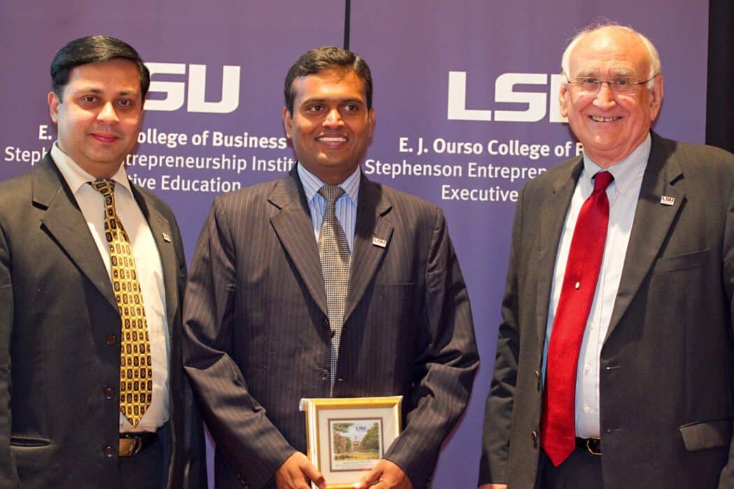 Three men in business attire stand in front of LSU banners; the middle man holds a framed award and all are smiling at the camera.