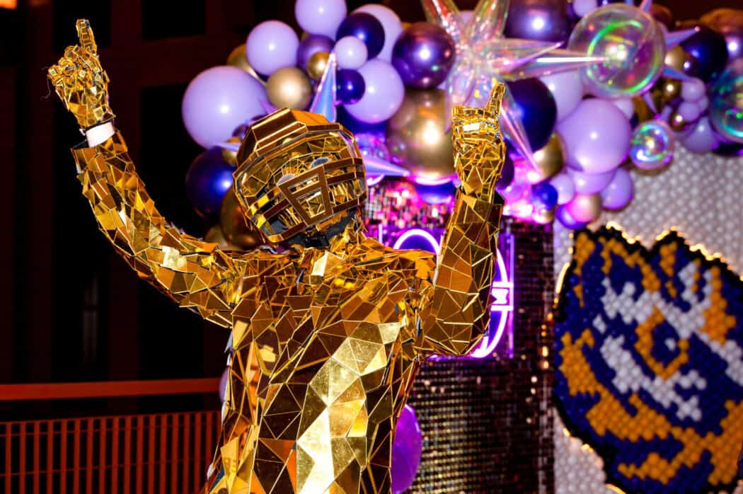 A person in a reflective gold mirror suit and helmet poses with both hands up; purple and gold balloons decorate the background.