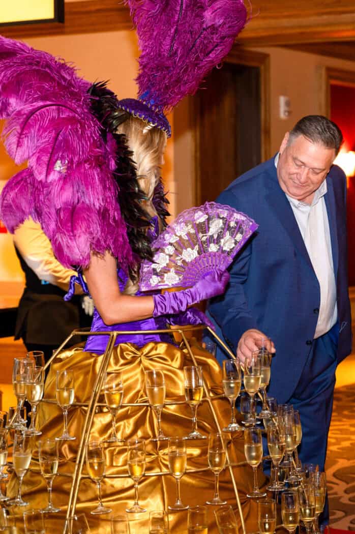 A man in a blue suit takes a glass of champagne from a tray attached to a performer dressed in a gold and purple costume with large feathers.