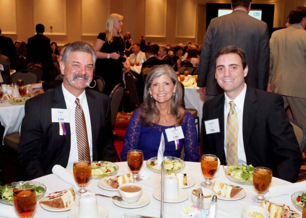 Three people in formal attire sit at a round table set with salads, tea, and plates at a crowded banquet event.