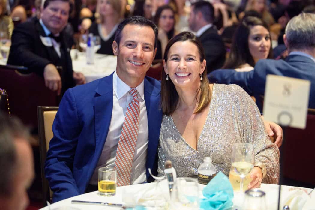 A man in a blue suit and a woman in a sparkly dress smile while seated at a round table at a formal event with other guests.