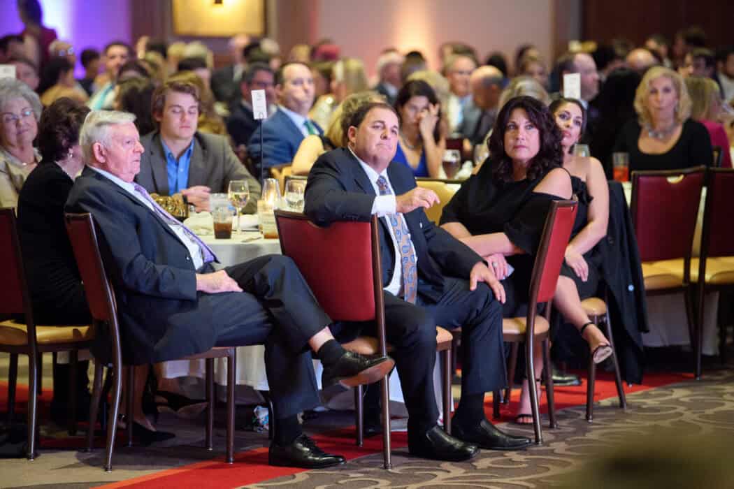 A group of people in formal attire sit at round tables watching an event or presentation in a large banquet hall.