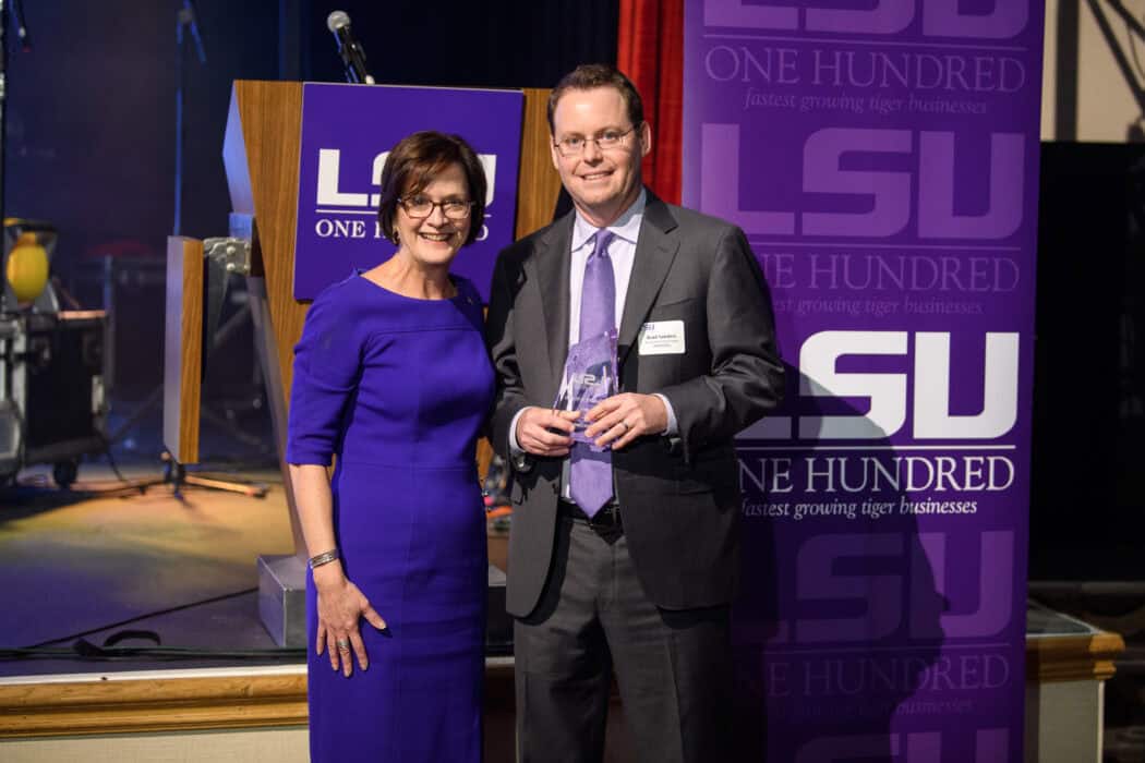 Two people stand together at an LSU ONE HUNDRED event; one holds a glass award, and both are dressed in formal attire.