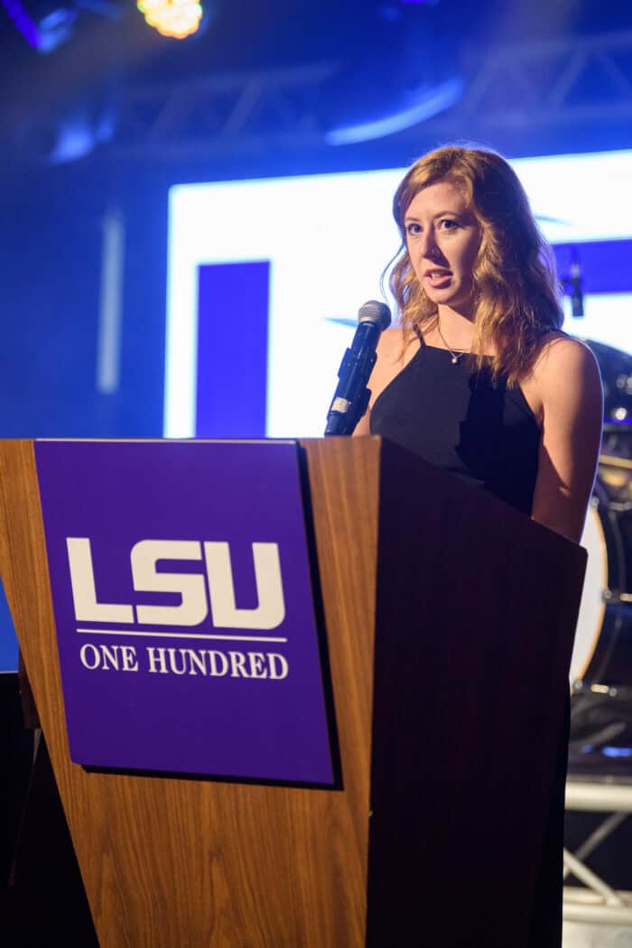 A woman in a black dress speaks at a podium with an LSU One Hundred sign, under stage lights.
