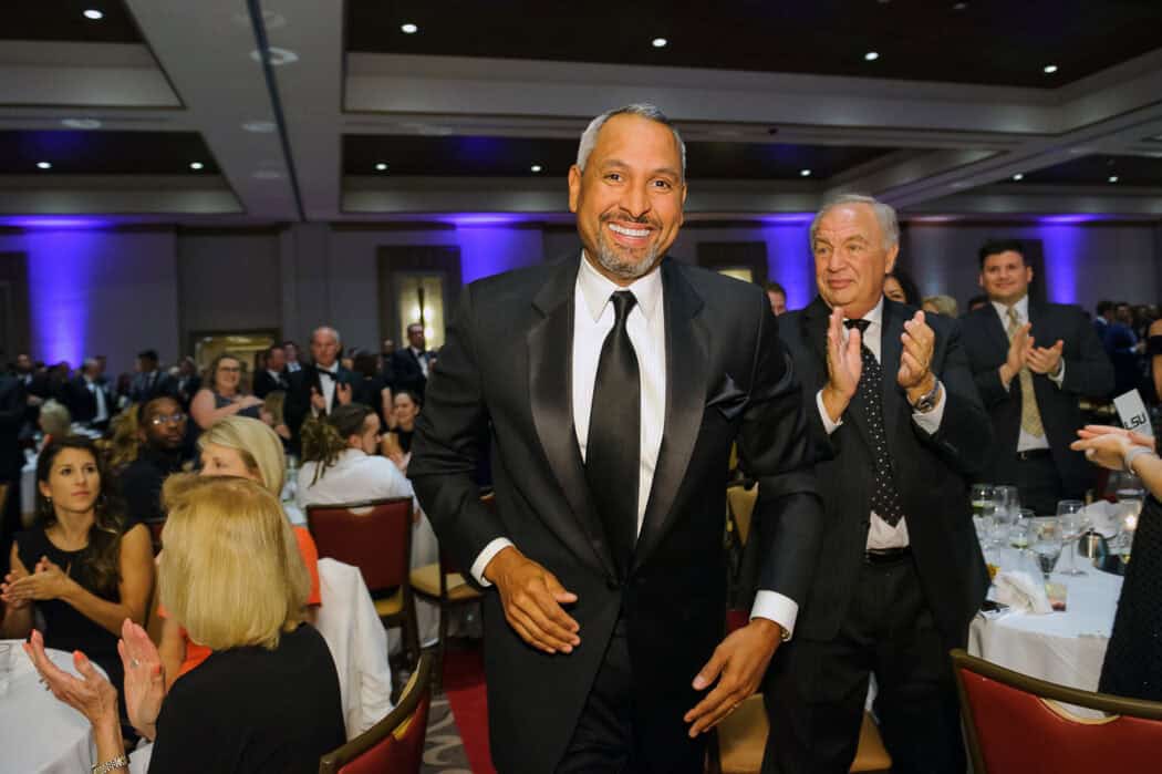 A man in a black suit smiles as he walks through a crowd at a formal event; people around him are seated and clapping.