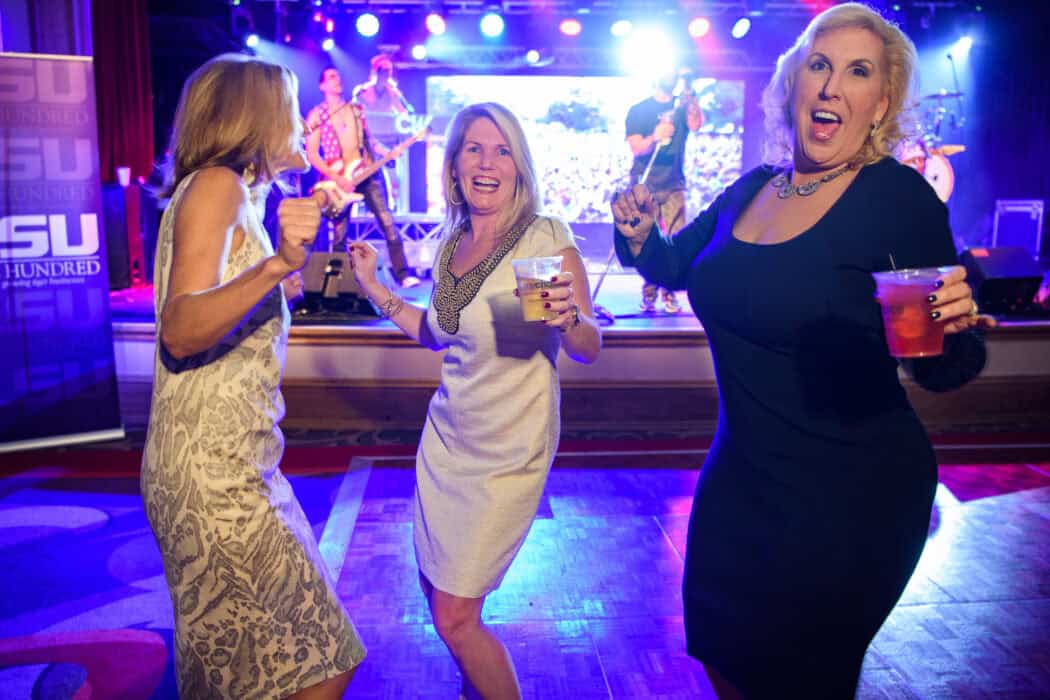 Three women in dresses are dancing and holding drinks at an indoor event with a live band playing in the background.