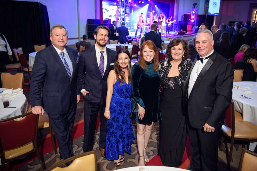 Six people in formal attire pose together in a banquet hall with a stage and live band performing in the background.
