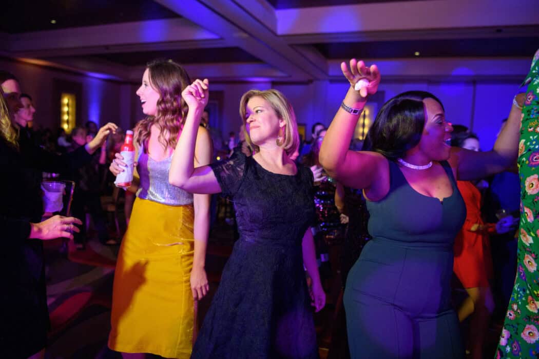 A group of people in formal attire dance together in a room with colorful, purple stage lighting.