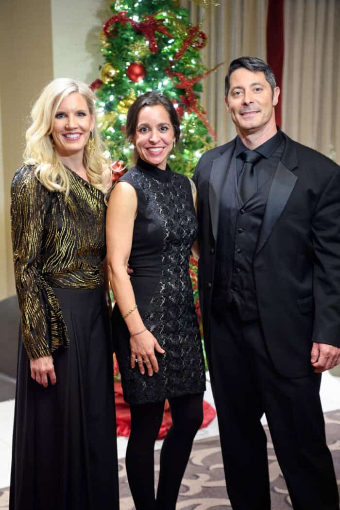 Three adults dressed in formal attire stand together and smile in front of a decorated Christmas tree.