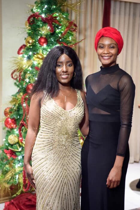 Two women stand in front of a decorated Christmas tree, one in a gold dress and the other in a black dress with a red headwrap.