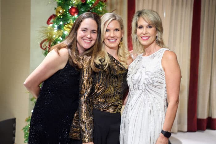 Three women in formal dresses stand together and smile in front of a decorated Christmas tree and beige curtains.