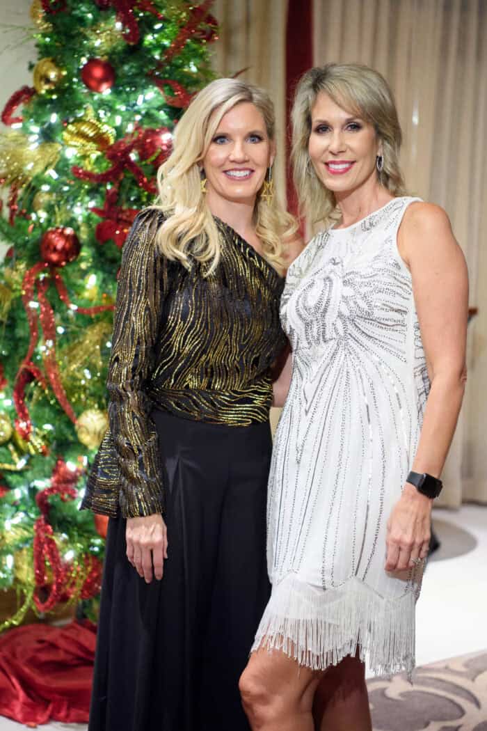 Two women in festive dresses stand and smile in front of a decorated Christmas tree with red and gold ornaments.