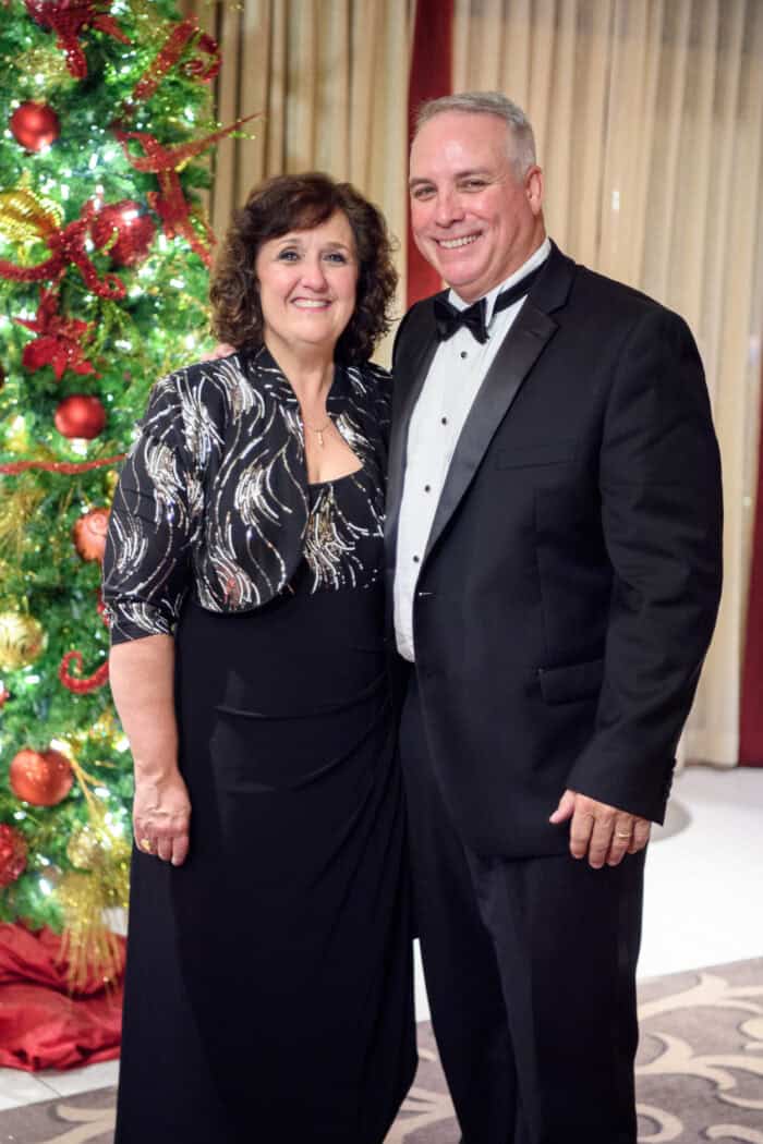 A woman and a man in formal attire stand smiling beside a decorated Christmas tree indoors.