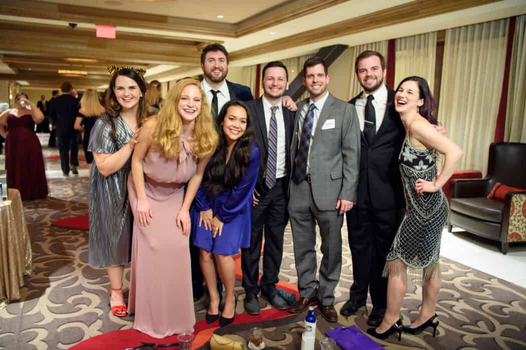 Eight adults dressed in formal wear smile and pose together at an indoor event with other guests in the background.
