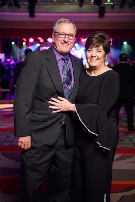 A man in a suit and a woman in a black dress pose together and smile at an indoor event with colorful lights in the background.
