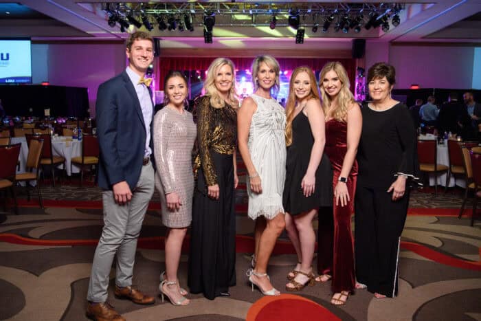 Seven people, dressed in formal attire, stand in a row and pose for a group photo at an indoor event with tables and stage in background.