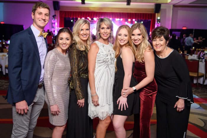 Seven people dressed in formal attire stand together posing for a group photo at an indoor event with colorful lights in the background.