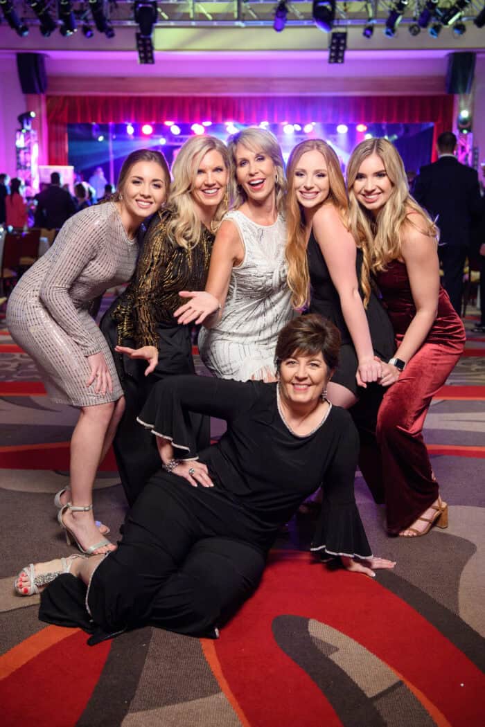 Six women pose and smile together at an indoor event with colorful stage lights and patterned carpet in the background.