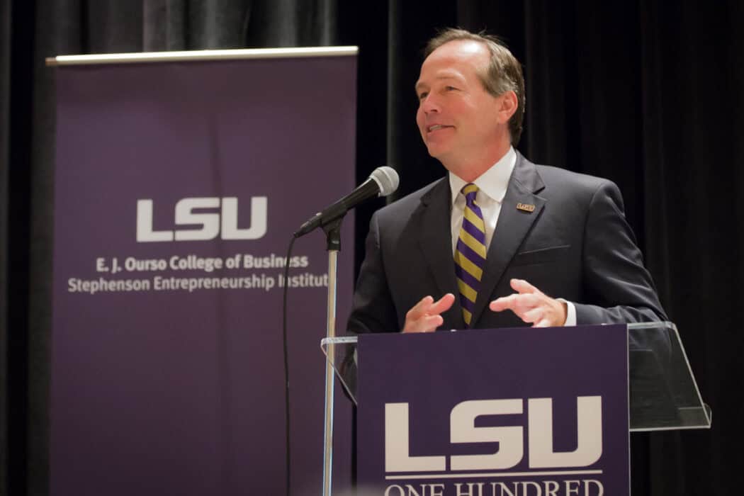 A man in a suit speaks at a podium with LSU logos and signage in the background at an event.