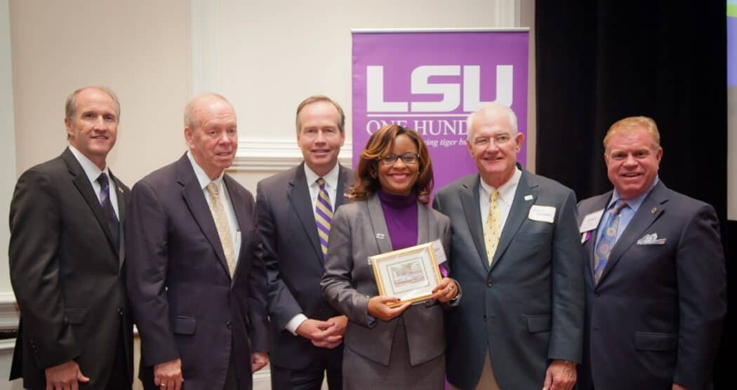 Six people in business attire stand in a row; the woman in the center holds a framed certificate. An LSU banner is visible behind them.