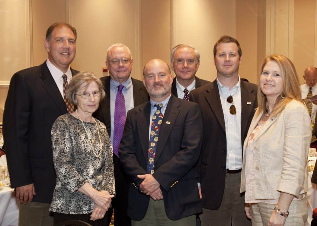 Seven adults, four men in suits, one man in a tie, and two women, stand together indoors, posing for a group photo at an event.