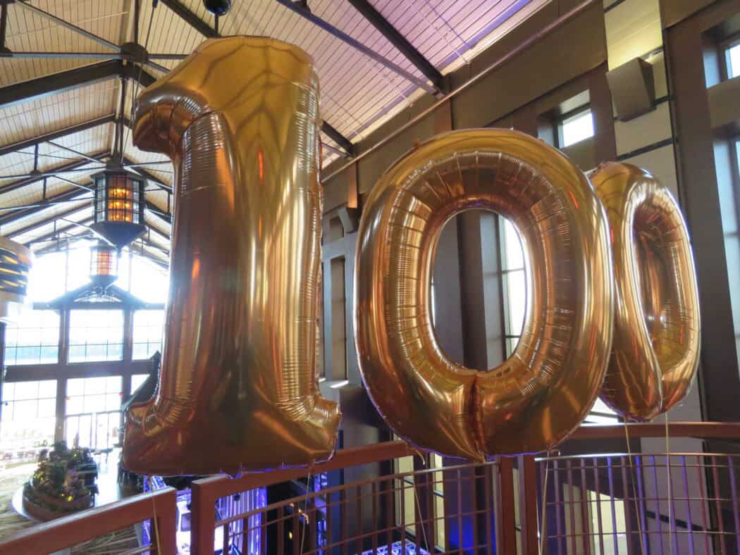 Large gold balloons in the shape of the number 100 are displayed indoors near a railing in a spacious, well-lit building.