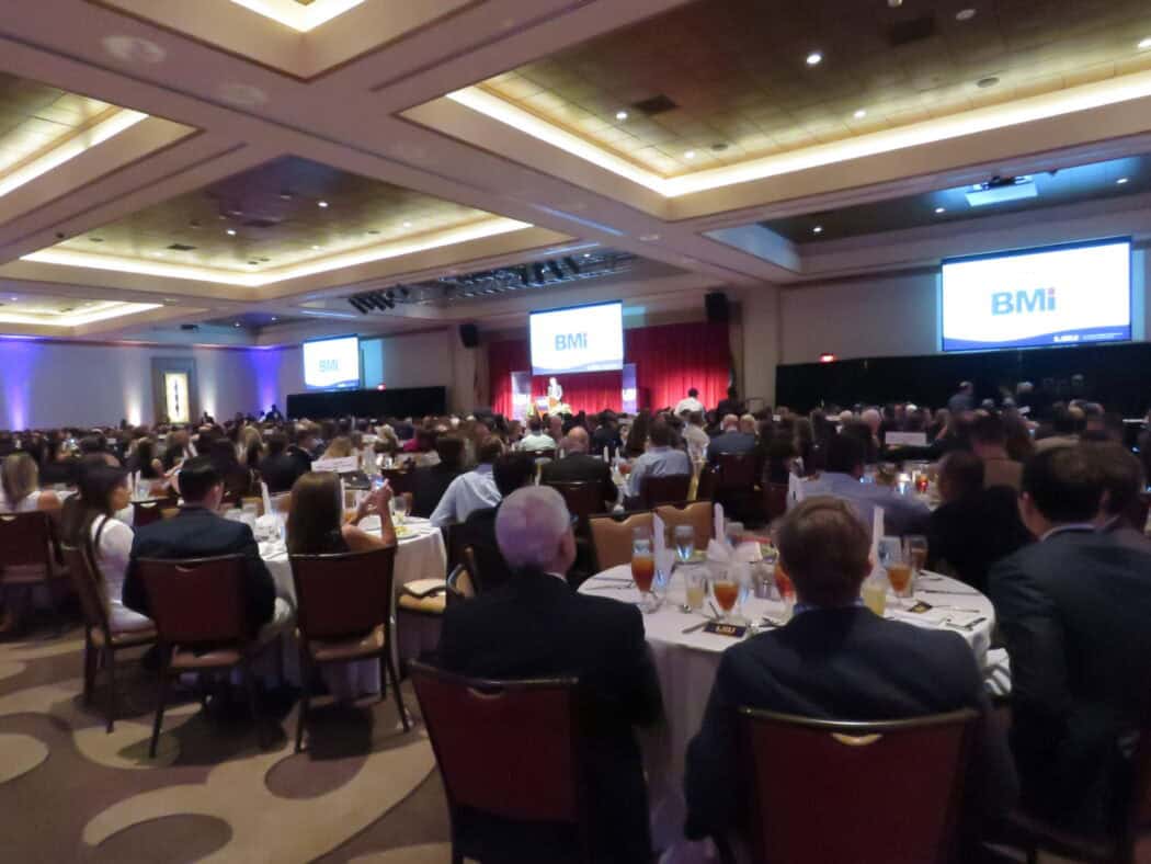 A large group of people seated at round tables in a banquet hall, watching a speaker on stage with "BMi" displayed on screens.