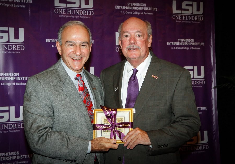 Two men in suits pose, one holding a framed item with a ribbon, in front of an LSU One Hundred event backdrop.