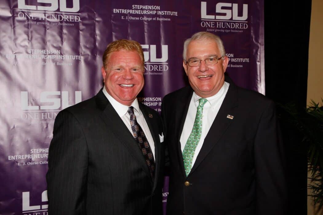 Two men in suits smile for a photo in front of a purple LSU One Hundred event backdrop.