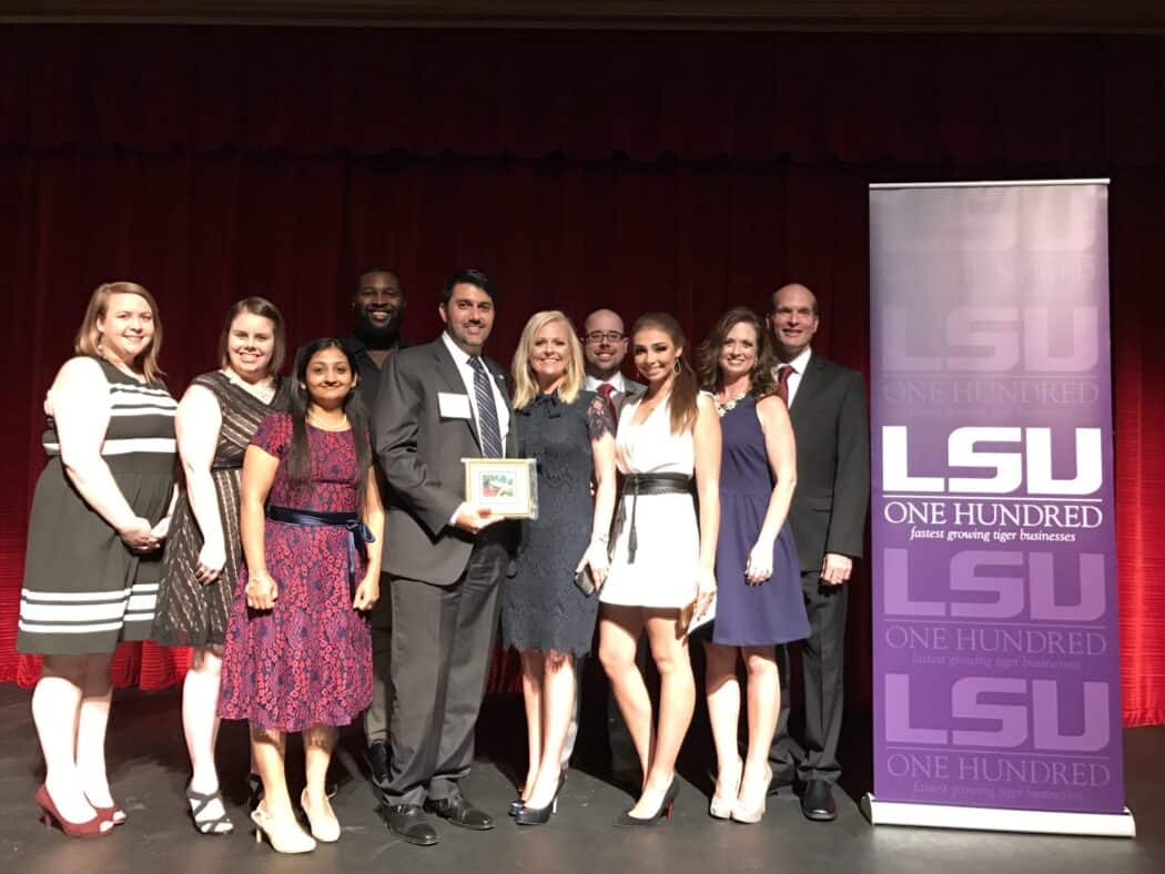 A group of people pose on stage next to an LSU ONE HUNDRED banner; one person holds a framed award.